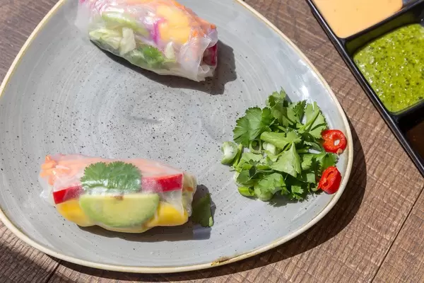 Nahaufnahme von Rainbow Sommerrollen mit Avocado, Mango, Paprika, Rotkohl, Karotten und Reisnudeln serviert mit Koriander und bunten Saucen