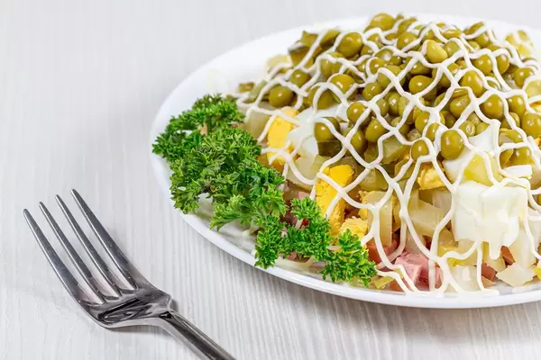 Nahaufnahme von russischem Oliviersalat aus Eiern, Kartoffeln, Erbsen, Fleisch und Petersilien auf weißem Teller