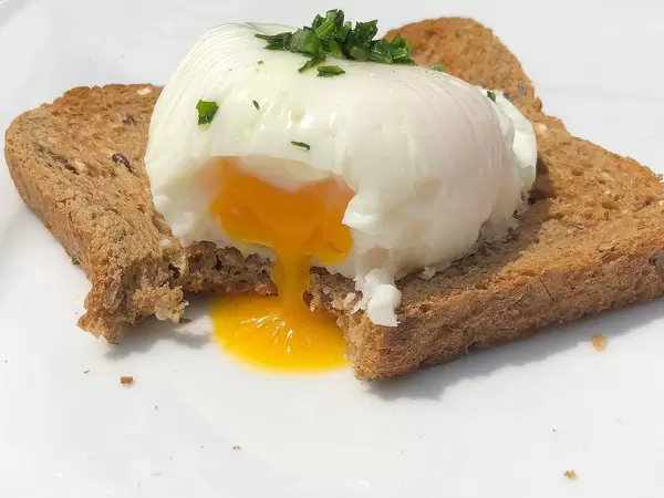 Nahaufnahme zeigt ein angebissenes pochiertes Ei auf Vollkornbrot, mit flüssigem Eigelb, auf einem weißen Tisch, als vegetarische Frühstücksidee