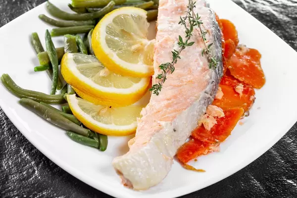 Nahaufnahme zeigt ein fertiges Stück Lachs mit Gemüse, Thymian und Zitronenscheiben auf einem weißen Teller, mit schwarzer Schiefersteinplatte im Hintergrund