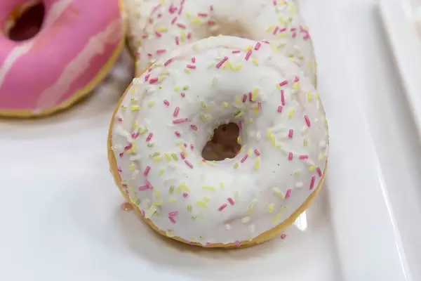 Nahaufnahme zeigt einen Donut mit weißer Glasur und pinken Streuseln, auf einem weißen Tablett