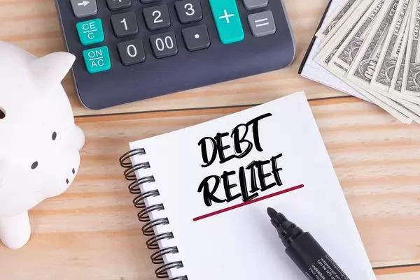 Notizbuch mit Text Debt Relief, Sparschwein, Taschenrechner und Dollar Geldscheine auf einem Holztisch