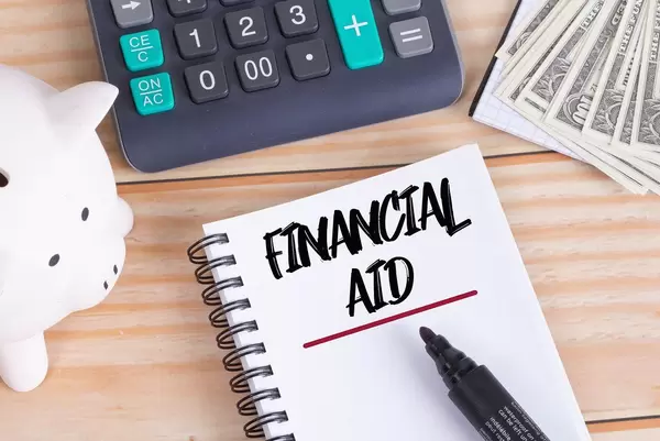 Notizbuch mit Text Financial Aid, Sparschwein, Taschenrechner und Dollar Geldscheine auf einem Holztisch