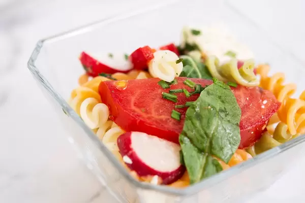Nudelsalat mit Tomate, Radieschen, Rucola und Käse in Glasschale vor weißem Hintergrund