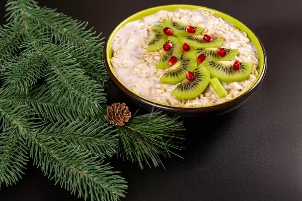 Oat flakes with kiwi, pomegranate and yogurt on a black background with branches of a christmas tree