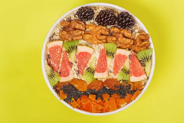 Oatmeal in a white bowl with blackberries, walnuts, grapefruit, kiwi and dried apricots, yellow background, top view