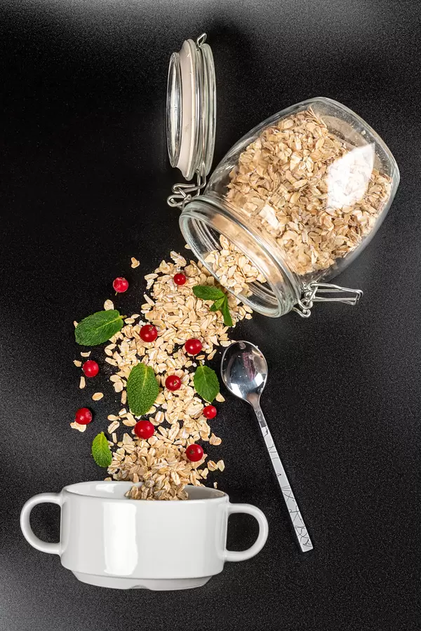 Oatmeal poured from a jar with red currant berries and mint on a black background with a white bowl and spoon