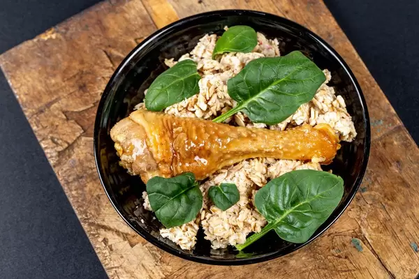 Oatmeal with baked chicken leg and green leaves on an old wooden kitchen board, top view (Flip 2020)
