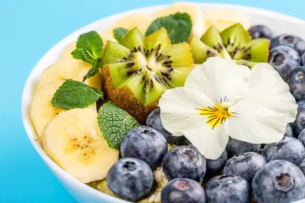 Oatmeal with blueberry, banana, mint, kiwi and white flower on blue background, close-up