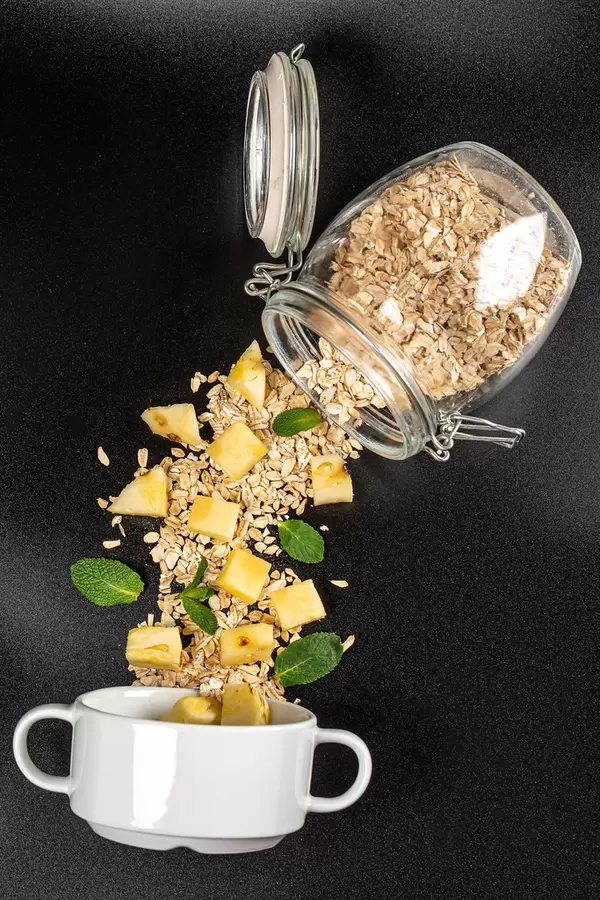 Oatmeal with pineapple pieces and mint leaves poured from a jar on a dark background with a white bowl, top view