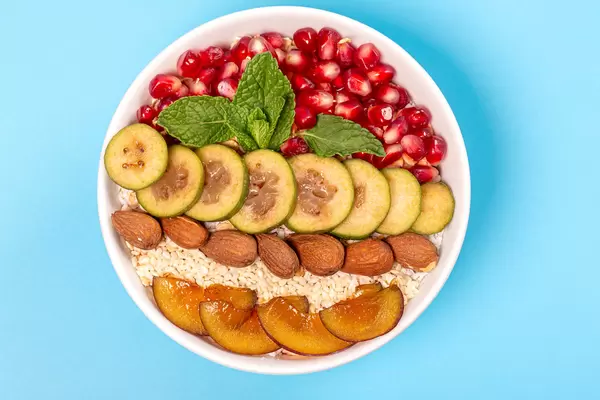 Oatmeal with pomegranate, pieces of feijoa, plum, almonds and mint leaves on a blue background, top view