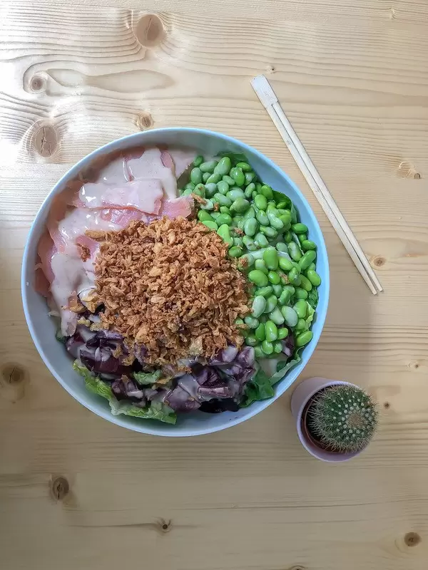 Obene Aufnahme vom Salmon Bowl mit Lachs, rote Beete, Edamame, Röstzwiebeln und Soja-Sesam Soße mit Essstäbchen und ein Kaktus auf dem Tisch