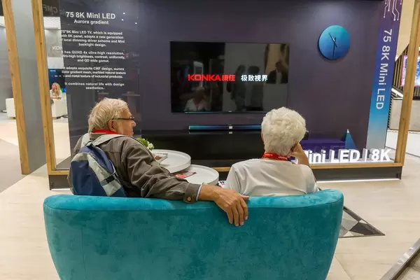 Oma und Opa auf der Couch: Altes Pärchen sitzt beim Messebesuch auf dem Sofa, vor einem 75 8K Mini LED Fernseher von Konka