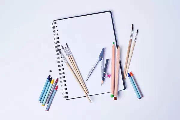 Open notepad with empty page and group of color pencils, brushes and compass