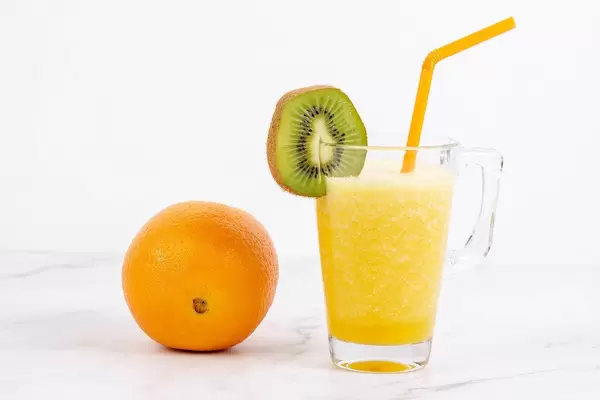 Orangesaft in Glas, dekoriert mit Trinkhalm und Kiwi-Scheibe neben ganzer Orange vor weiß