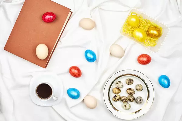 Ostern Hintergrund: gefärbte Eier, Wachteleier, Teller und Kaffeetasse und ein Buch auf weißem Bettlaken