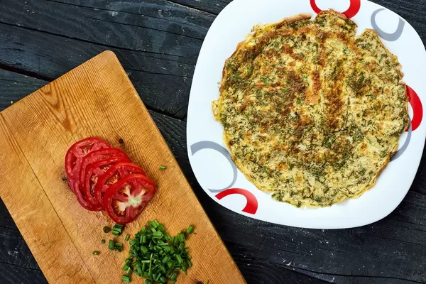 Overhead shot of herbs based omelet and wooden cutting board with slices of tomato and scallions