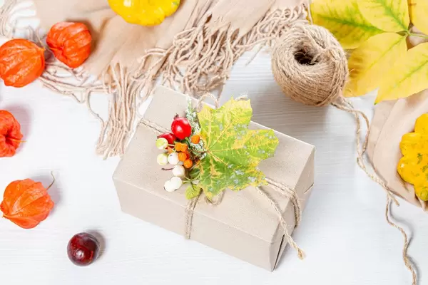 Packaged gift with yellow leaves, berries, scarf and thread on white wooden table. Family holiday concept, Thanksgiving Day (Flip 2019)