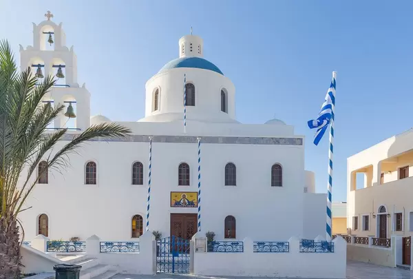 Panagia Platsani Kirche in Oia, Santorin mit blauer Kuppel, Kirchenglocken und griechischer Flagge