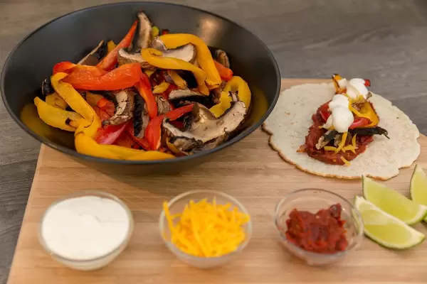 Paprikastreifen mit Pilzen in einer schwarzen Schüssel und geriebener Chedderkäse, Tomatenmark, Limettenschmand mit einer Fajita auf einem Brettchen