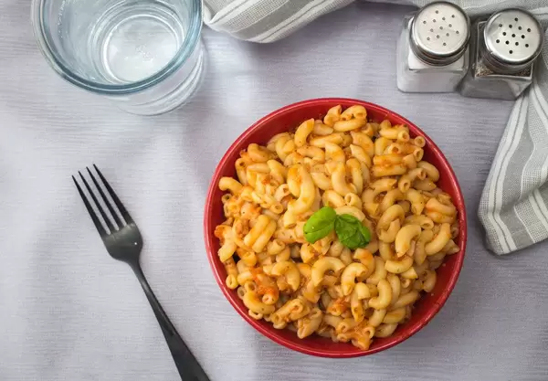 Pasta - Nudeln mit Tomatensoße und Basilikum in einer Roten Schale in der Aufsicht auf einem Tisch mit Gabel, einem Glas Wasser und einem Salzstreuer