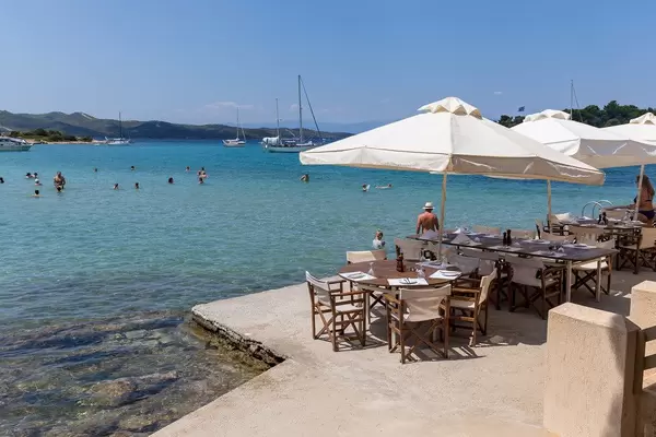 People swimming in the blue sea at Hinitsa Beach, next to restaurant tables under parasols, on a greek island
