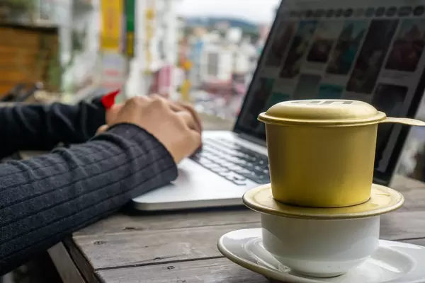 Person arbeitet online am Laptop mit Vietnamesischem Filterkaffee im Vordergrund Nahaufnahme