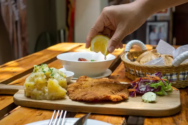 Person drückt eine Zitrone über einem Hähnchen Schnitzel aus auf einem Holzbrett mit Kartoffelsalat, Beilagensalat und Kräuterbutter in einem deutschen Restaurant