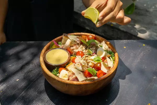 Person drückt eine Limette über einem gesundem Salat mit Hähnchenbrust, Parmesan, Kirschtomaten, Basilikum, Geröstetem Brot und Honig Senf Sauce aus