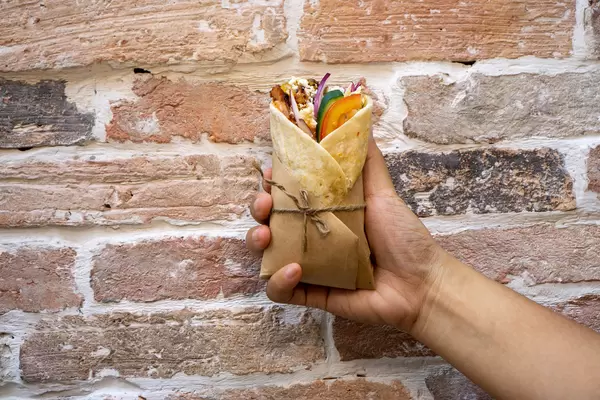 Person hält einen Mini Döner Dürum Wrap mit Hänchenfleisch, Tomaten, Zwiebeln, Gurken, Rotkohl, Tzatziki und Feta Käse in der Hand vor einer Ziegelwand