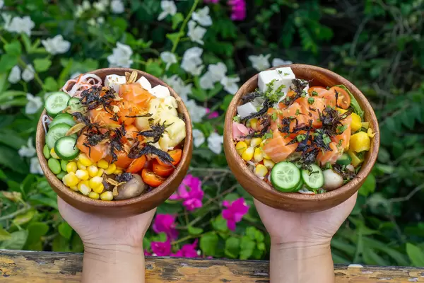 Person hält zwei Holzschalen mit verschiedenen frischen Zutaten als Hawaiianischen Poke Bowl in den Händen mit Blumen im Hintergrund