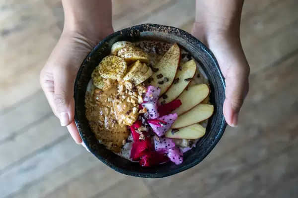 Person hält ein gesundes Porridge mit Apfel, Banane, Pitaya, Zimt, Kokosnussmilch und Erdnussbutter in der Hand