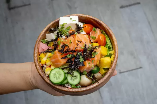 Person hält eine Holzschüssel mit Gesunder Hawaiianischer Poke Bowl mit verschiedenem rohem Gemüse, Lachs, Tofu, getrocknetem Seetang Draufsicht