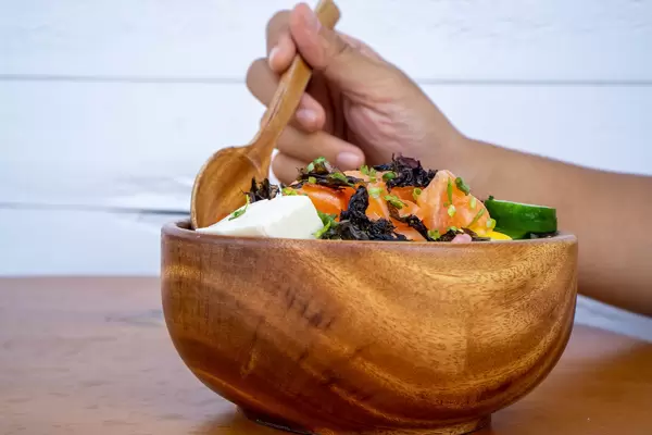 Person isst eine gesunde Hawaiianische Poke Bowl mit Tofu, rohem Lachs und Gemüse mit einem Holzlöffel Nahaufnahme