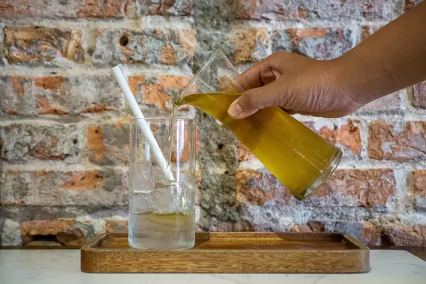 Person pouring Iced Green Tea Small Glass Pitcher in a Drinking Glass with Ice Cubes and Paper Straw