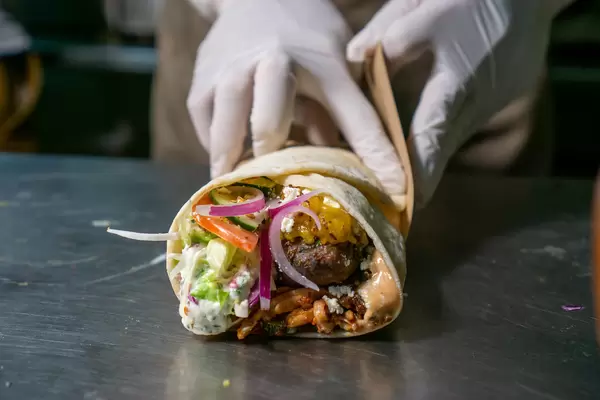 Person rollt einen Döner Dürum Wrap mit gegrilltem Lammfleisch, frischem Gemüse, Käse, Tzatziki und Cocktail Soße auf einer Edelstahl Arbeitsplatte Nahaufnahme