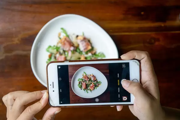Person taking Food Photo with a Smartphone of Honeydew Melon wrapped in Parma Ham with Arugula, Parmesan Ham and Balsamic Dressing on a Ceramic Plate