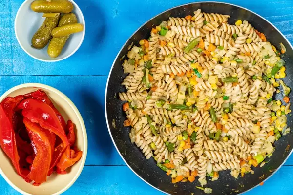 Pfanne mit gebratenen Fusilli-Nudeln mit Gemüse neben Schalen mit Essiggurken und roter Paprika auf einem blauen Holztisch Draufsicht