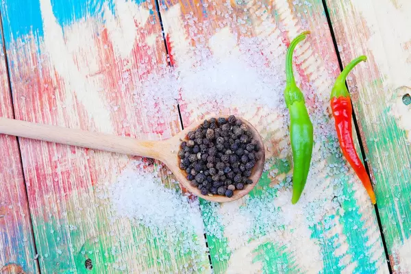 Pfefferkörner auf einem Holzlöffel mit Salz und zwei Chilis auf buntem Hintergrund