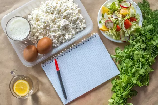 Planning diet concept. Flat lay view of notepad with a pen and organic healthy food: cottage cheese, eggs, greens, salad, apple, and greens on brown paper (Flip 2019)