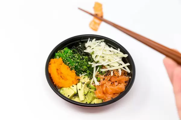 Poke Bowl Lachs Teriyaki - mit Sushireis, Lachs, Avocado, Krautsalat, Wakame Salat, Wasago, Nori, Teriyaki-Sauce, Soja-Sauce, Sesam und Schnittlauch - Ein Stück Lachs wird mit den Essstäbchen genommen