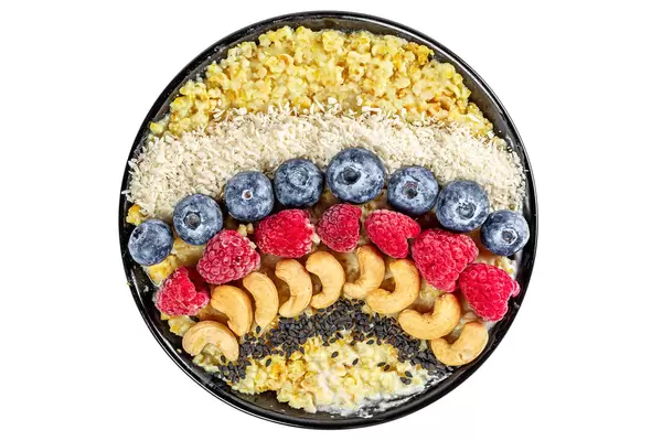 Porridge for breakfast with black sesame seeds, cashews, raspberries, blueberries and coconut on a white background, top view
