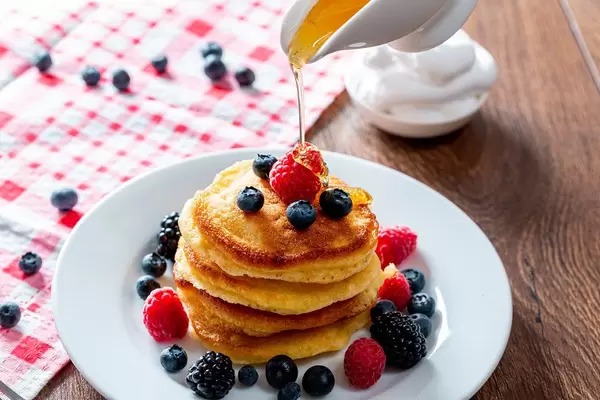 Pouring honey onto tasty pancakes with berries on table, closeup