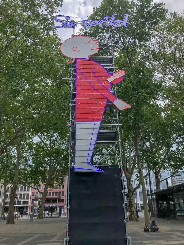 Provokantes Kunstwerk & Streetart von Alexandra Berlinger & Martin Wagner am Drogentreffpunkt Neumarkt in Köln, zeigt ein Reisdorf-Männchen beim "Schuss setzen" und dem Slogan: Sie spritzt