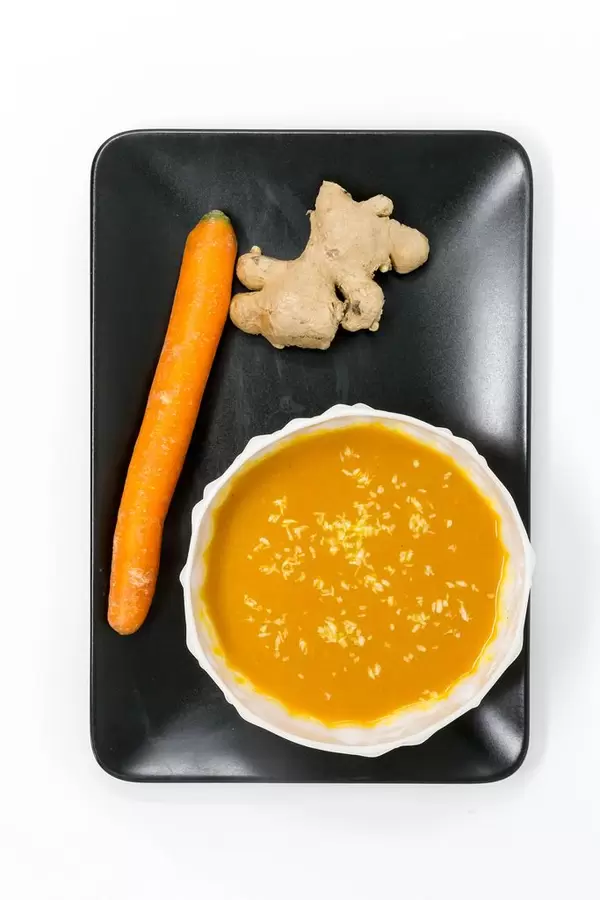 Pumpkin Coconutsoup with Ginger in a bowl placed on a black plate with a carrot and a ginger root