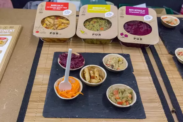 PUR - veganer Bio Nudelsalat, Quinoasalat und Rote Betesalat als Proben in Schälchen mit Verpackungen auf einer Messer