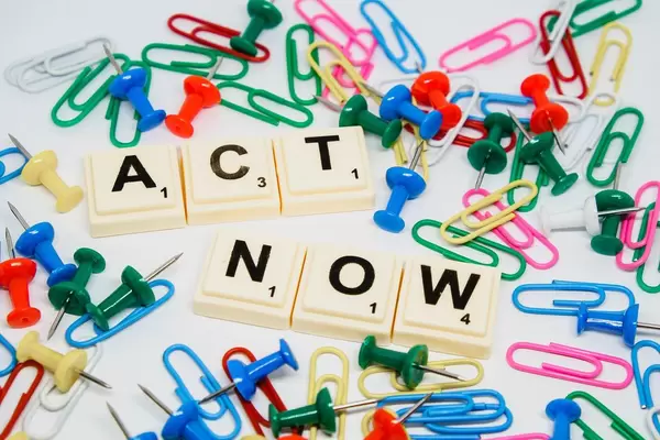 "Act Now" -Handel jetzt Schriftzug mit Scrabblesteinen, umgeben von bunten Reißzwecken und Büroklammern