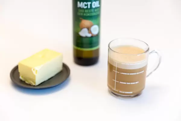 "Bullet Proof Coffee" in a glass cup, in front of a bottle of coconut MCT oil by Woldo Health and a piece of butter, is supposed to create a feeling of satiety during a diet