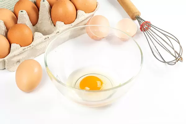 Raw egg in glass bowl on white background with eggs in tray and whisk
