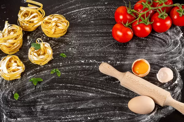 Raw fettuccine, tomatoes, eggs and flour with rolling pin on black background